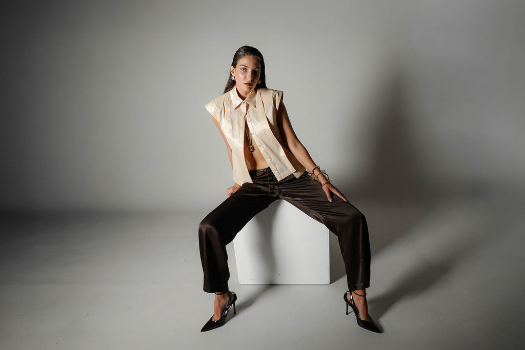 Landscape campaign shot of the Realm Origin Sol Verite Shirt in powder beige, structured sleeveless taffeta shirt with front flange pleats and tailored shoulders, styled on-model in an editorial seated pose.