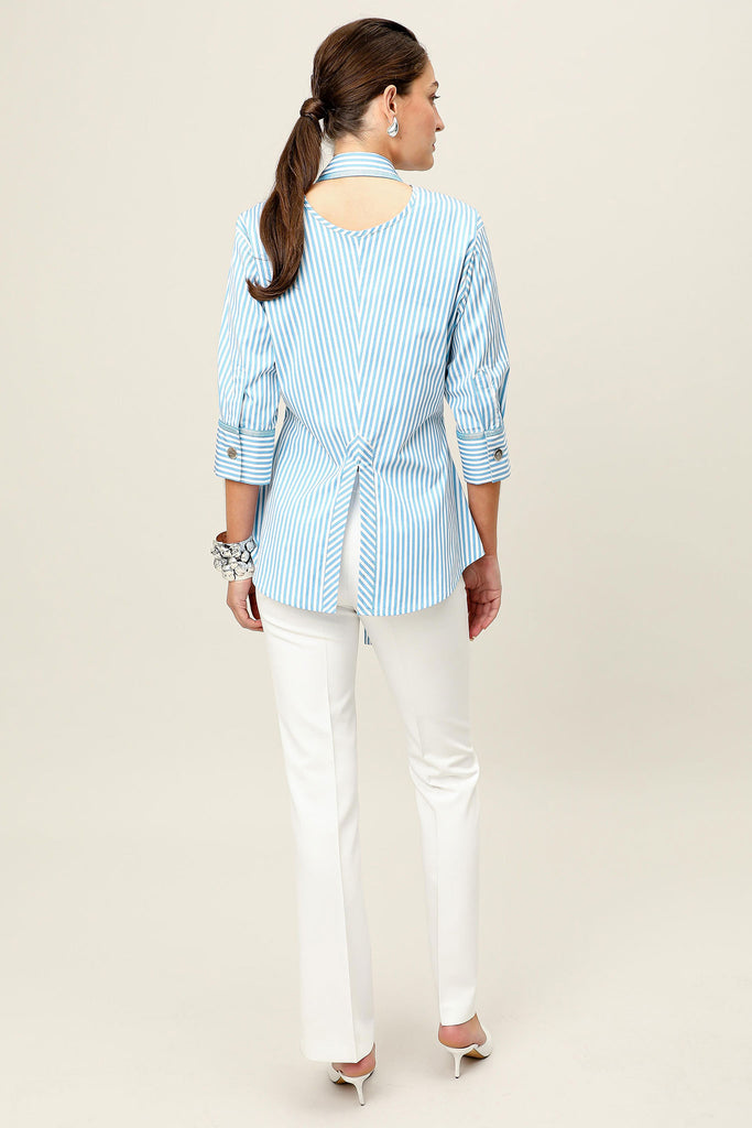 Model wearing the Vera ova blue and white striped tailored shirt by Realm Origin, shown from the back highlighting the pleated detail and cut-out open collar, paired with white trousers and heels against a neutral background.