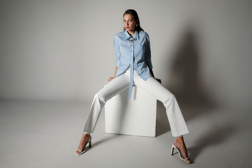 Model wearing the Vera Nova blue and white striped tailored shirt by Realm Origin, styled with white trousers and heels, sitting on a white cube in a studio setting with soft shadows.
