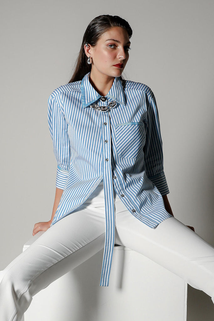 Model wearing the Vera Nova blue and white striped tailored shirt by Realm Origin, featuring a pointed collar, long front placket detail, paired with white trousers, seated on a stool against a neutral background.