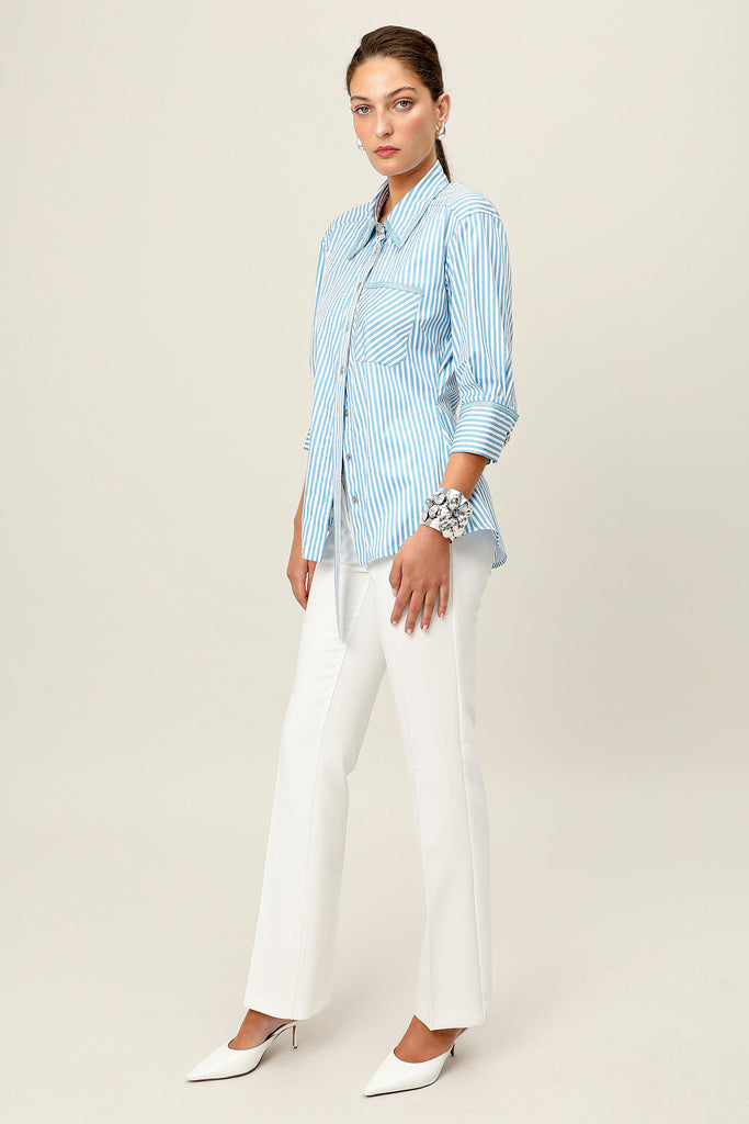 Model wearing the Vera Nova blue and white striped tailored shirt by Realm Origin, shown from a side angle, styled with white trousers and heels against a neutral background.